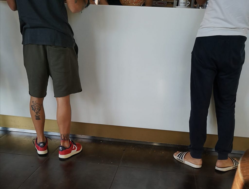Italienische Männer an der Bar, es sind nur de Beine zu sehen mit Adiletten und Sportschuhen